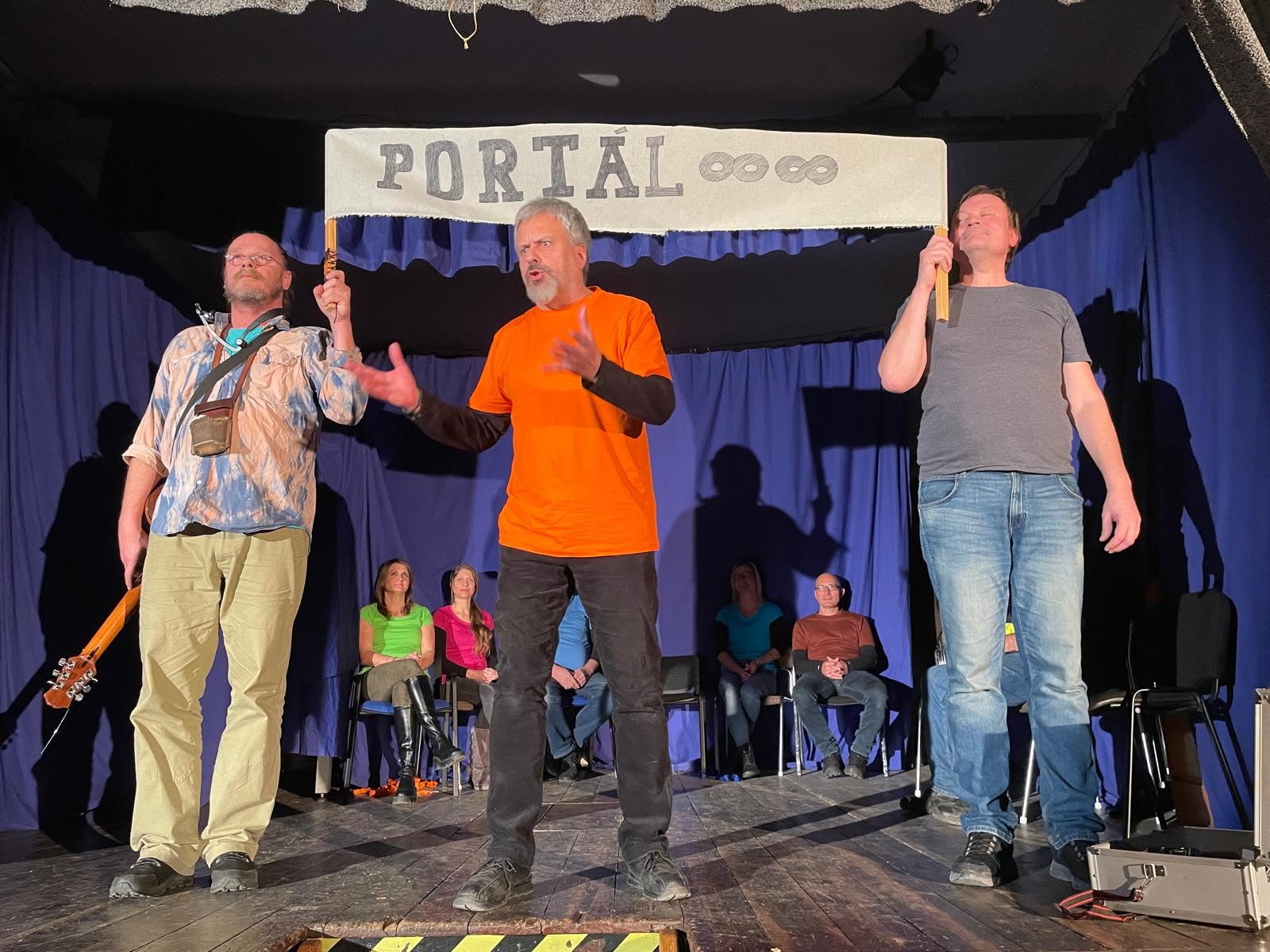 Tři herci na jevišti pod cedulí s nápisem Portál